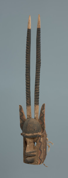 rzeźba , maska - Ujęcie z przodu skosem w lewą stronę. Część twarzowa: czoło wypukłe, czarne, cienki grzbiet nosa, szerokie nozdrza, stożkowate usta. wycięte trójkątne oczy. Na obu bokach części twarzowej wymalowany zyg-zakowaty wzór (czarna linia, wypełnienie trójkątów - czerwone). Część twarzowa zwieńczona lekko wygiętymi do tyłu długimi żłobkowanymi rogami oraz szpiczastymi uszami ozdobionymi zyg-zakowatymi wzorami (kolory analogicznie jak wyżej). W części tylnej maski wywiercone otwory, umożliwiające przymocowanie jej do twarzy tancerza oraz pozostałości po sznurku.
