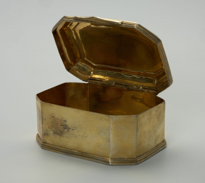 pozłacane puzderko na niskim, profilowanym cokoliku - Ujęcie z przodu, z szeroko uchyloną pokrywą; Pozłacane puzderko na niskim, profilowanym cokoliku. Korpus ośmioboczny, na rzucie prostokąta ze ściętymi narożami. Pokrywa na zawiasie, z dwoma uskokami, lekko wybrzuszona z wielokrotnie profilowanym brzegiem, z przodu w centralnej części wysunięty wydłużony płaski uchwyt w formie listwy o zaoblonych narożach, trójkątnym wykroju i z delikatnym profilowaniem. Powierzchnia puzderka na zewnątrz i we wnętrzu złocona, poza częścią spodnią.