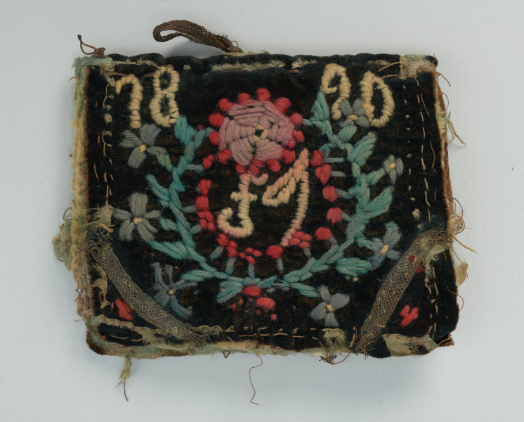 książeczka na igły z haftowanymi okładkami i kartkami z wełny - Ujęcie z przodu; Przedmiot w kształcie książeczki z czarną, materiałową okładką i czterema kartkami z miękkiej, wełnianej tkaniny w kolorze naturalnym, służących do wbijania igieł. Całość zszyta razem, brzegi obszyte atłasową taśmą w zielonym kolorze. Przód  zdobi haft płaski, wykonany włóczką w kolorach czerwonym, żółtym i zielonym. Haft przedstawia wieniec, w środku którego widnieje monogram 
