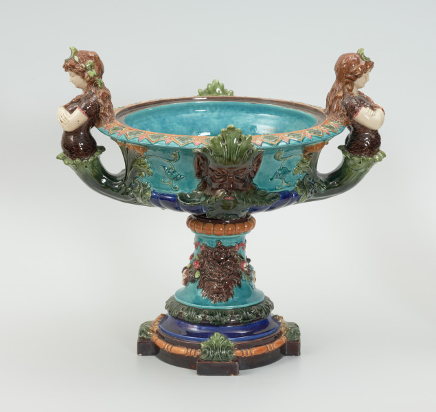 Waza - Ujęcie z przodu. Waza w kształcie smukłej amfory. Brzusiec ceramiczny, rozszerzający się ku górze, w kolorze kremowym, malowany podszkliwnie (z uzupełnieniami naszkliwnymi) we wzory kwiatowe - bukiety barwnych kwiatów w typie aster w kolorach fioletowo-czerwonym, liliowym, żółtym i białym, z gałązkami i listkami oraz drobnymi kwiatkami przypominającymi bratki o barwie fioletowo-czerwonej i liliowej. Górna część wazy wraz z uchwytami oraz stopą i część podstawy wykonane jako odlewy z mosiądzu. Mosiężna szyja wąska z otokiem perełkowym w środku ozdobiona jest pionowym, rozszerzającym się ku dołowi i ku górze rytmicznym podziałem z ornamentem geometryczno-roślinnym. Na pogrubionym skraju szyi umieszczona jest płytka miseczka w formie kwiatu o wąskich płatkach, stanowiąca wylewkę wazonu. Poniżej dolnej części szyi oprawa wierzchołka brzuśca składająca się z otoku o sercowym wycięciu zwieńczonym profilowaną palmetką oraz dwóch wydłużonych, sięgających 1/3 wysokości brzuśca, pionowych wsporników uchwytów. Te ostatnie składają się z 2 części: zewnętrzna wychodząca ze wsporników opartych na brzuścu, wyniesiona jest po niewielkim skosie do połowy wysokości szyi i na końcu wywinięta do wnętrza wolutowo, a druga - krótsza, skierowana ku dołowi łączy łagodnym łukiem wolutę z górną częścią oprawy brzuśca, kończąc się wywiniętymi na zewnątrz listkami. Uchwyty żłobkowane, zdobione motywem liści. Wazon wsparty jest na mosiężnej podstawie, składającej się z wieńca oplecionego wstęgą, na którym spoczywa zwężająca się ku górze stopa o brzegach ozdobionych perełkowaniem i pierścieniem, wspierającym koszyczek, ujmujący dolną część ceramicznego brzuśca, w formie kwiatu o ostro zakończonych, wąskich płatkach. Całość spoczywa na podstawie z kwadratowej płytki czerwonego marmuru ze ściętymi narożnikami, ujętymi mosiężnymi wspornikami ramy wspierającej płytę od dołu i opartej w narożach na czterech lwich łapach.