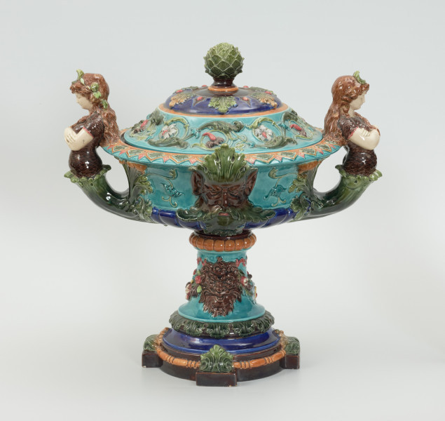 Waza - Ujęcie z tyłu z pokrywką. Waza w kształcie smukłej amfory. Brzusiec ceramiczny, rozszerzający się ku górze, w kolorze kremowym, malowany podszkliwnie (z uzupełnieniami naszkliwnymi) we wzory kwiatowe - bukiety barwnych kwiatów w typie aster w kolorach fioletowo-czerwonym, liliowym, żółtym i białym, z gałązkami i listkami oraz drobnymi kwiatkami przypominającymi bratki o barwie fioletowo-czerwonej i liliowej. Górna część wazy wraz z uchwytami oraz stopą i część podstawy wykonane jako odlewy z mosiądzu. Mosiężna szyja wąska z otokiem perełkowym w środku ozdobiona jest pionowym, rozszerzającym się ku dołowi i ku górze rytmicznym podziałem z ornamentem geometryczno-roślinnym. Na pogrubionym skraju szyi umieszczona jest płytka miseczka w formie kwiatu o wąskich płatkach, stanowiąca wylewkę wazonu. Poniżej dolnej części szyi oprawa wierzchołka brzuśca składająca się z otoku o sercowym wycięciu zwieńczonym profilowaną palmetką oraz dwóch wydłużonych, sięgających 1/3 wysokości brzuśca, pionowych wsporników uchwytów. Te ostatnie składają się z 2 części: zewnętrzna wychodząca ze wsporników opartych na brzuścu, wyniesiona jest po niewielkim skosie do połowy wysokości szyi i na końcu wywinięta do wnętrza wolutowo, a druga - krótsza, skierowana ku dołowi łączy łagodnym łukiem wolutę z górną częścią oprawy brzuśca, kończąc się wywiniętymi na zewnątrz listkami. Uchwyty żłobkowane, zdobione motywem liści. Wazon wsparty jest na mosiężnej podstawie, składającej się z wieńca oplecionego wstęgą, na którym spoczywa zwężająca się ku górze stopa o brzegach ozdobionych perełkowaniem i pierścieniem, wspierającym koszyczek, ujmujący dolną część ceramicznego brzuśca, w formie kwiatu o ostro zakończonych, wąskich płatkach. Całość spoczywa na podstawie z kwadratowej płytki czerwonego marmuru ze ściętymi narożnikami, ujętymi mosiężnymi wspornikami ramy wspierającej płytę od dołu i opartej w narożach na czterech lwich łapach.