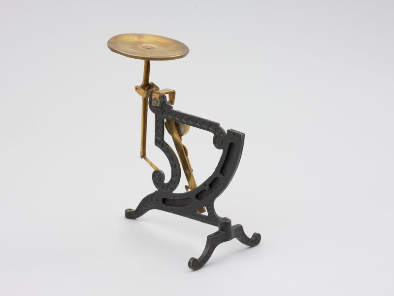 jednowahadłowa uchylna waga pocztowa - Ujęcie z tyłu lewoboczne; Jednowahadłowa uchylna waga pocztowa. WPodstawa wagi składa się z dwóch elementów, z których element poprzeczny przymocowany jest do głównego elementu na ruchomo, co pozwala składać nóżkę poprzez jej przekręcenie. Dłuższe ramię podstawy zakończone jest śrubką pozwalającą na regulację wagi w opozycji pionowej. Stalowa konstrukcja łącząca główny trzon wagi z dźwignią nośną nasadzona jest na szalkę.