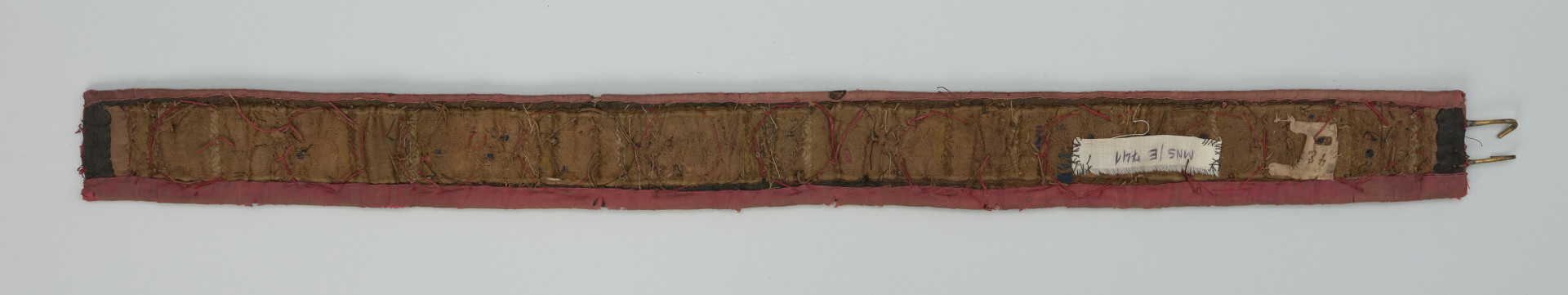 Tkany weselny pas kobiecy - Ujęcie z tyłu. Wąski pasek obciągnięty od spodu brązową tkaniną atłasową. Z prawej strony odchodzą od niego dwa zaczepy w postaci hakowato wygiętych na końcach pręcików.