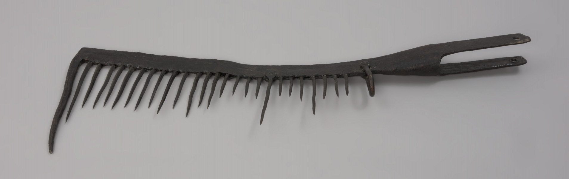 grzebień - Ujęcie z góry. 26-zębna ość grzebieniasta, wykonana techniką kowalską. Ramie robocze płaskie, z jednego końca zagięte pod kątem 90 stopni (część ta ma formę zęba grubszego i dłuższego od pozostałych), w drugim końcu przechodzi w tuleję z dwoma pasami, służącą do osadzenia narzędzia na trzonku (trzonka brak). Na końcu każdego pasa otwór – jednego okrągły, drugiego – kwadratowy. Przy nasadzie tulei uszko wykonane z wygiętego w kształcie litery „U” pręta o okrągłym przekroju. Mocowane za pomocą nitu. Zęby mocowane w ramieniu, nierównej długości, ostro zakończone.  Ząb ósmy, licząc od uszka, przynitowany, długi.