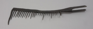 grzebień - Ujęcie z góry. 26-zębna ość grzebieniasta, wykonana techniką kowalską. Ramie robocze płaskie, z jednego końca zagięte pod kątem 90 stopni (część ta ma formę zęba grubszego i dłuższego od pozostałych), w drugim końcu przechodzi w tuleję z dwoma pasami, służącą do osadzenia narzędzia na trzonku (trzonka brak). Na końcu każdego pasa otwór – jednego okrągły, drugiego – kwadratowy. Przy nasadzie tulei uszko wykonane z wygiętego w kształcie litery „U” pręta o okrągłym przekroju. Mocowane za pomocą nitu. Zęby mocowane w ramieniu, nierównej długości, ostro zakończone.  Ząb ósmy, licząc od uszka, przynitowany, długi.