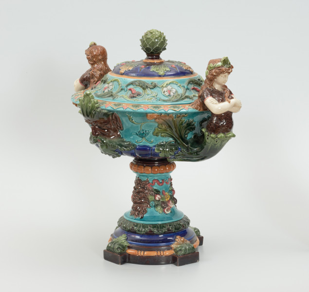 Waza - Ujęcie z przodu skosem w lewą stronę. z pokrywką. Waza w kształcie smukłej amfory. Brzusiec ceramiczny, rozszerzający się ku górze, w kolorze kremowym, malowany podszkliwnie (z uzupełnieniami naszkliwnymi) we wzory kwiatowe - bukiety barwnych kwiatów w typie aster w kolorach fioletowo-czerwonym, liliowym, żółtym i białym, z gałązkami i listkami oraz drobnymi kwiatkami przypominającymi bratki o barwie fioletowo-czerwonej i liliowej. Górna część wazy wraz z uchwytami oraz stopą i część podstawy wykonane jako odlewy z mosiądzu. Mosiężna szyja wąska z otokiem perełkowym w środku ozdobiona jest pionowym, rozszerzającym się ku dołowi i ku górze rytmicznym podziałem z ornamentem geometryczno-roślinnym. Na pogrubionym skraju szyi umieszczona jest płytka miseczka w formie kwiatu o wąskich płatkach, stanowiąca wylewkę wazonu. Poniżej dolnej części szyi oprawa wierzchołka brzuśca składająca się z otoku o sercowym wycięciu zwieńczonym profilowaną palmetką oraz dwóch wydłużonych, sięgających 1/3 wysokości brzuśca, pionowych wsporników uchwytów. Te ostatnie składają się z 2 części: zewnętrzna wychodząca ze wsporników opartych na brzuścu, wyniesiona jest po niewielkim skosie do połowy wysokości szyi i na końcu wywinięta do wnętrza wolutowo, a druga - krótsza, skierowana ku dołowi łączy łagodnym łukiem wolutę z górną częścią oprawy brzuśca, kończąc się wywiniętymi na zewnątrz listkami. Uchwyty żłobkowane, zdobione motywem liści. Wazon wsparty jest na mosiężnej podstawie, składającej się z wieńca oplecionego wstęgą, na którym spoczywa zwężająca się ku górze stopa o brzegach ozdobionych perełkowaniem i pierścieniem, wspierającym koszyczek, ujmujący dolną część ceramicznego brzuśca, w formie kwiatu o ostro zakończonych, wąskich płatkach. Całość spoczywa na podstawie z kwadratowej płytki czerwonego marmuru ze ściętymi narożnikami, ujętymi mosiężnymi wspornikami ramy wspierającej płytę od dołu i opartej w narożach na czterech lwich łapach.