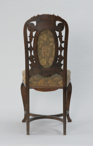 krzesło - Ujęcie z tyłu. Krzesło w stylu neobarokowym, oparcie i siedzisko tapicerowane. Nogi zwężające się do dołu, przednie wygięte kabriolowo, górne wybrzuszenie ozdobione płaskorzeźbionym motywem palmety, zakończenie dolne trójkątne. Tylne nogi lekko wygięte, powiązane są z przednimi krzyżową poprzeczką umieszczoną u dołu, składającą się z dwóch stykających się w środku półokrągłych pałąkowych listew. Konstrukcja zaplecka stanowi pionowe przedłużenie tylnych nóg o profilowanych krawędziach, zamykających się dwoma półkołami w kształt serca, ponad którym wyprowadzono łukowe zwieńczenie w formie mięsiście rzeźbionego, rozłożonego na boki bukietu liści. Poniżej sercowego styku listew bocznych wydłużony w pionie medalion opasany płaską listwą z wypełnieniem tapicerką. Tkanina w kwiatowy wzór w odcieniach zieleni, różu, żółcieni i ciemnego popielu. Pomiędzy owalem medalionu i listwami oparcia ażurowe wypełnienie mięsistą płaskorzeźbą o motywach roślinnych. Taką samą tkaniną obite jest siedzisko o konstrukcji sprężynowej, stosunkowo wysokie, poszerzające się do przodu, w dolnej części wykończone półwałkiem i pasmanterią, kryjącą krawędź tapicerki.