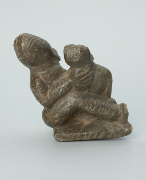 figura kultu przodków - Ujęcie z boku z prawej. Rzeźbione w grafitowym kamieniu dwie postaci ludzkie w pozycji siedzącej. Mniejsza postać – najprawdopodobniej dziecko - siedzi na kolanach większej - najprawdopodobniej kobiety. Obie postacie ozdobiono licznymi nacięciami liniowymi. Widoczne rysy i mikropęknięcia.