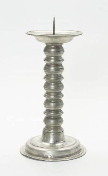 cynowy świecznik na okrągłej, wysklepionej stopie - Ujęcie z przodu; Cynowy świecznik ołtarzowy z trzonem balaskowym, na okrągłej, wysklepionej stopie, z profitką w formie talerzyka i iglicą do mocowania świecy. Stopa ozdobiona na skraju wypłaszczenia tłoczonym rowkiem, zakończona jest dołem szerokim kręgiem podstawy, tworzącym kryzę z podwójnym tłoczonym rowkiem. Górna część stopy wyniesiona cylindrycznie tworzy oparcie trzonu, złożonego z nakładanych na siebie naprzemiennie pierścieni o dwóch formach: pięciu spłaszczonych kul o obłym wykroju oraz czterech (+ połówka na stopie) nieco niższych o wykroju spiczastym; w połowie wysokości pierścienie zdobi półwałek, zaś górną i dolną nasadę tłoczony rowek. Profitka z szeroką, płaską kryzą ma średnicę zbliżoną do wymiaru stopy.