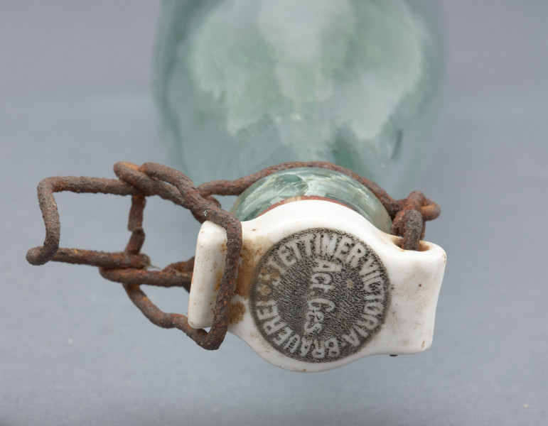 szklana butelka z kapslem - detal; Kapsel z białego porcelitu owalny z wypustami na dwóch bokach. W centrum kapsla okrągły znak na ciemnoszarym tle, po owalu napis: STETTINER VICTORIA BRAUEREI