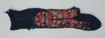 Wełniana pończocha z kwiatowym haftem - Ujęcie z przodu; Pończocha wykonana z granatowej wełny gęstym ściegiem pończoszniczym. Zdobiona haftem płaskim. Wzór wyszyty na stopie, po obu stronach kostek , na łydce. Haft o motywach roślinnych w postaci dużych kwiatów czerwono - zielonych. Pomiędzy nimi sieć drobnych listków wypełniających gęsto pole zdobnicze. Kompozycję od góry zamyka umieszczony centralnie, duży kwiat, wokół którego znajduje się rozeta złożona z pięciu kwiatów.