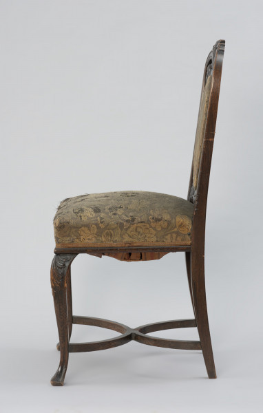 krzesło - Ujęcie z prawego boku. Krzesło w stylu neobarokowym, oparcie i siedzisko tapicerowane. Nogi zwężające się do dołu, przednie wygięte kabriolowo, górne wybrzuszenie ozdobione płaskorzeźbionym motywem palmety, zakończenie dolne trójkątne. Tylne nogi lekko wygięte, powiązane są z przednimi krzyżową poprzeczką umieszczoną u dołu, składającą się z dwóch stykających się w środku półokrągłych pałąkowych listew. Konstrukcja zaplecka stanowi pionowe przedłużenie tylnych nóg o profilowanych krawędziach, zamykających się dwoma półkołami w kształt serca, ponad którym wyprowadzono łukowe zwieńczenie w formie mięsiście rzeźbionego, rozłożonego na boki bukietu liści. Poniżej sercowego styku listew bocznych wydłużony w pionie medalion opasany płaską listwą z wypełnieniem tapicerką. Tkanina w kwiatowy wzór w odcieniach zieleni, różu, żółcieni i ciemnego popielu. Pomiędzy owalem medalionu i listwami oparcia ażurowe wypełnienie mięsistą płaskorzeźbą o motywach roślinnych. Taką samą tkaniną obite jest siedzisko o konstrukcji sprężynowej, stosunkowo wysokie, poszerzające się do przodu, w dolnej części wykończone półwałkiem i pasmanterią, kryjącą krawędź tapicerki.