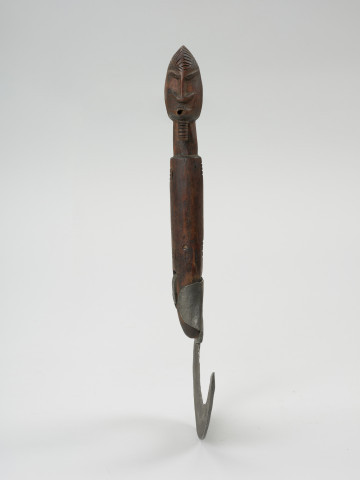 narzędzie rolnicze - Ujęcie z przodu. Sierp o kutym z żelaza, łukowatym ostrzu. Rączka drewniana, walcowata, zakończona rzeźbą główki z brodą w formie nacinanego liniami walca.