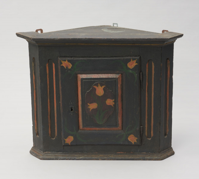 meble - Ujęcie z przodu; Szafka kątowa z płycinowymi, jednoskrzydłowymi drzwiami i dekoracją z motywem tulipanów. Konstrukcja skrzyniowa oparta na planie trójkąta ze ściętymi narożnikami. Gzyms prosty olistwowany, dół obwiedziony profilowaną listwą. Drzwi płycinowe, jednoskrzydłowe z zamkiem i ozdobną wykładką, w środku półki. Płycina dekorowana wypukłym, owalnym emblematem z namalowanymi trzema tulipanami. Na narożnikach drzwi także namalowane tulipany. Ściana licowa i boczne dekorowane wklęsłymi, podłużnymi polami o lekko zaokrąglonych końcach. Całość ciemnogranatowa, motywy kwiatowe w kolorach pomarańczowym i zielonym. Ozdobne linie na ścianach i gzymsie zaznaczone na pomarańczowo.