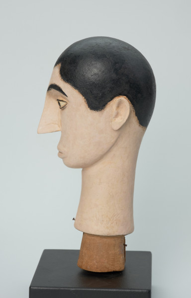 lalka teatralna: Tubabu Cè Kun - Ujęcie z lewego boku. Drewniane popiersie mężczyzny. Głowa owalna, zwężająca się ku dołowi. Zaznaczone krótkie włosy z falistą grzywką, pomalowane na czarno. Twarz i szyja pomalowane na blado-różowy, cielisty kolor. Zaznaczone czarne, łukowate brwi, migdałowe, wyrzeźbione oczy z powiekami i czarnym cienkim obramowaniem, z czarną okrągłą źrenicą w środku. Nos wąski, długi, usta lekko wypukłe, pomalowane na kolor cielisty. Szyja wąska, długa, rozszerzająca się ku dołowi.