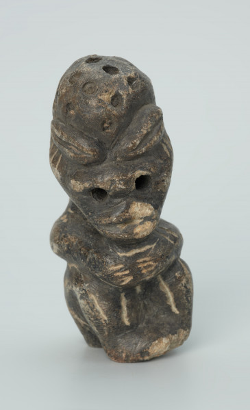 rzeźba; przedmiot obrzędowy; Figura kultu zmarłych - Ujęcie z przodu z prawej strony. Rzeźbiona w grafitowobeżowym steatycie siedząca postać ludzka, której nieproporcjonalnie dużą głowę zdobią liczne, okrągłe wgłębienia, natomiast ciało pokrywają pionowe i ukośne nacięcia, oznaczające prawdopodobnie skaryfikację. Powierzchnia rzeźby z rysami, mikropęknięciami, przetarciami oraz drobnymi uszkodzeniami.