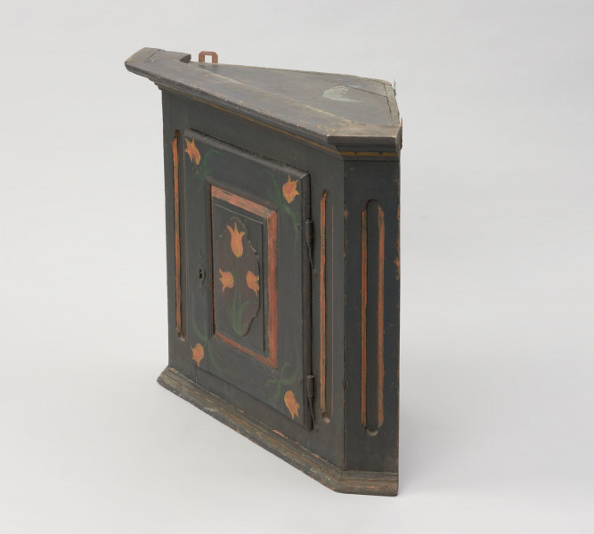 meble - Ujęcie przodu i drugiego boku; Szafka kątowa z płycinowymi, jednoskrzydłowymi drzwiami i dekoracją z motywem tulipanów. Konstrukcja skrzyniowa oparta na planie trójkąta ze ściętymi narożnikami. Gzyms prosty olistwowany, dół obwiedziony profilowaną listwą. Drzwi płycinowe, jednoskrzydłowe z zamkiem i ozdobną wykładką, w środku półki. Płycina dekorowana wypukłym, owalnym emblematem z namalowanymi trzema tulipanami. Na narożnikach drzwi także namalowane tulipany. Ściana licowa i boczne dekorowane wklęsłymi, podłużnymi polami o lekko zaokrąglonych końcach. Całość ciemnogranatowa, motywy kwiatowe w kolorach pomarańczowym i zielonym. Ozdobne linie na ścianach i gzymsie zaznaczone na pomarańczowo.