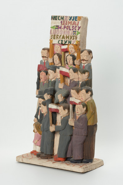 rzeźba - Ujęcie z przodu skosem z lewej strony. Rzeźba wykonana w jednym kawałku drewna lipowego . Opracowana tylko od strony frontalnej. Wolnostojąca na prostokątnym postumencie jest , bryłą zwartą, słabo rozczłonkowaną, statyczną. Postacie umieszczone wertykalnie: w trzech rzędach sami mężczyźni ubrani w garnitury, trzymający w rękach biało - czerwone i czerwoną chorągiewki. Powyżej kobiety ( widoczne w zasadzie same głowy) trzymają transparent z napisem: 