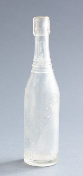 szklana butelka z wyodrębnioną szyjką z trzema pierścieniami - Ujęcie z przodu; Szklana butelka piwówka z grubego szkła butelkowego półbiałego przezroczystego. Szyjka wyodrębniona trzema wypukłymi, wąskimi pierścieniami, umieszczonymi w dolnej części szyjki. U dołu pogrubionego wylewu pogrubienia na dwa otwory do zamknięcia pałąkowego. Dno zagłębione płaskie. Zamknięcie pałąkowe. Ślad szwu bocznego.