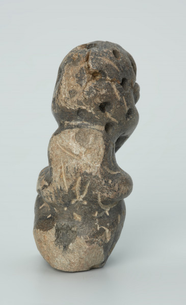 rzeźba; przedmiot obrzędowy; Figura kultu zmarłych - Ujęcie z tyłu. Rzeźbiona w grafitowobeżowym steatycie siedząca postać ludzka, której nieproporcjonalnie dużą głowę zdobią liczne, okrągłe wgłębienia, natomiast ciało pokrywają pionowe i ukośne nacięcia, oznaczające prawdopodobnie skaryfikację. Powierzchnia rzeźby z rysami, mikropęknięciami, przetarciami oraz drobnymi uszkodzeniami.