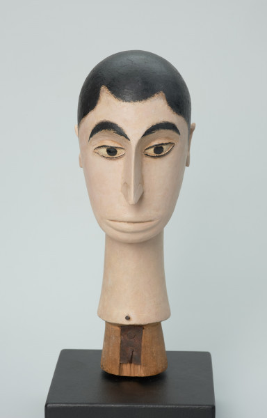 lalka teatralna: Tubabu Cè Kun - Ujęcie z przodu. Drewniane popiersie mężczyzny. Głowa owalna, zwężająca się ku dołowi. Zaznaczone krótkie włosy z falistą grzywką, pomalowane na czarno. Twarz i szyja pomalowane na blado-różowy, cielisty kolor. Zaznaczone czarne, łukowate brwi, migdałowe, wyrzeźbione oczy z powiekami i czarnym cienkim obramowaniem, z czarną okrągłą źrenicą w środku. Nos wąski, długi, usta lekko wypukłe, pomalowane na kolor cielisty. Szyja wąska, długa, rozszerzająca się ku dołowi.