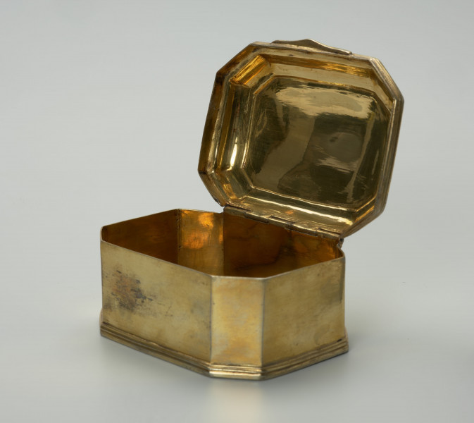 pozłacane puzderko na niskim, profilowanym cokoliku - Ujęcie z przodu, z bardzo szeroko uchyloną pokrywą; Pozłacane puzderko na niskim, profilowanym cokoliku. Korpus ośmioboczny, na rzucie prostokąta ze ściętymi narożami. Pokrywa na zawiasie, z dwoma uskokami, lekko wybrzuszona z wielokrotnie profilowanym brzegiem, z przodu w centralnej części wysunięty wydłużony płaski uchwyt w formie listwy o zaoblonych narożach, trójkątnym wykroju i z delikatnym profilowaniem. Powierzchnia puzderka na zewnątrz i we wnętrzu złocona, poza częścią spodnią.