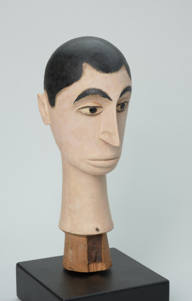 lalka teatralna: Tubabu Cè Kun - Ujęcie z przodu z prawej strony. Drewniane popiersie mężczyzny. Głowa owalna, zwężająca się ku dołowi. Zaznaczone krótkie włosy z falistą grzywką, pomalowane na czarno. Twarz i szyja pomalowane na blado-różowy, cielisty kolor. Zaznaczone czarne, łukowate brwi, migdałowe, wyrzeźbione oczy z powiekami i czarnym cienkim obramowaniem, z czarną okrągłą źrenicą w środku. Nos wąski, długi, usta lekko wypukłe, pomalowane na kolor cielisty. Szyja wąska, długa, rozszerzająca się ku dołowi.