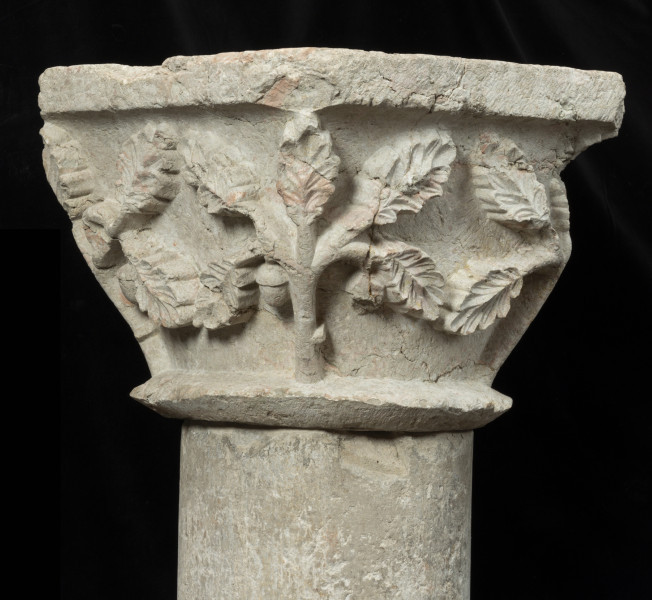 kapitel z przedstawieniem liści dębu - Ujęcie trzeciego boku. Kapitel blokowo-kielichowy, rozszerzający się ku górze, wykuty z jednego kawałka kamienia. Okrągłą podstawę otacza wałek o trójkątnym profilu. Zwieńczenie w formie szerokiej czworobocznej płyty abakusa. Płaskorzeźbioną powierzchnię między wałkiem i abakusem wypełniają ustawione pionowo w narożnikach pięciolistne gałązki dębu z pojedynczym żołędziem. Mięsiste liście ukazane frontalnie w układzie niemal symetrycznym. Wyraźnie zaznaczona falista linia brzegu blaszki i wklęsły ryt unerwienia liści. Relief półplastyczny, światłocieniowy. Tło opracowane gładko. Drobne pęknięcia na powierzchni kamienia. Liczne ukruszenia zwłaszcza w partii abakusa i dolnego walka. Pod spodem centralne wgłębienie do umocowania trzpienia łączącego kapitel z trzonem.