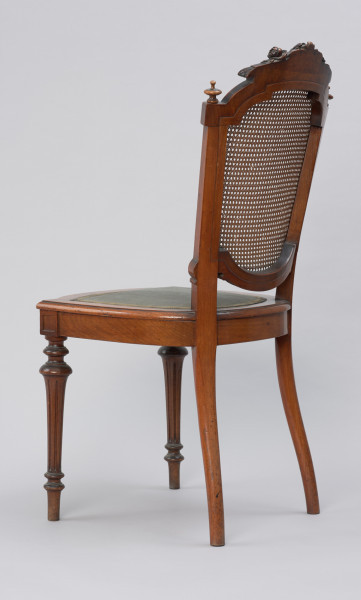 krzesło - Ujęcie z tyłu skosem w lewą stronę. Krzesło z płasko wyściełanym siedziskiem i oparciem z plecionki z rafii. Przednie nogi toczone, tralkowe, w części środkowej wydzielonej wałkami - kanelowane. Tylne nogi o przekroju okrągłym, lekko wygięte, przechodzące w prostokątne słupki zaplecka. Siedzenie w kształcie podkowiastym, z przodu lekko zaokrąglone na zewnątrz, w środku wyściełane - tkanina mocowana pinezkami tapicerskimi. Na styku przednich nóg i ramy siedziska ozdobna kostka z płycinami na obu ściankach zewnętrznych. Rama zaplecka w formie medalionu, wypełnionego plecionką z rafii. Słupki zaplecka dekorowane wgłębnym reliefem w formie płycin i spirali, a łuk zwieńczenia zdobi dekoracja o formach rollwerkowo-roślinnych. Ponad kostkami wieńczącymi boczne ramy zaplecka toczone gałki. Siedzisko wyściełane tkaniną w kolorze zielonym (turkusowym?), z brzegami wykończonymi pasmanterią.