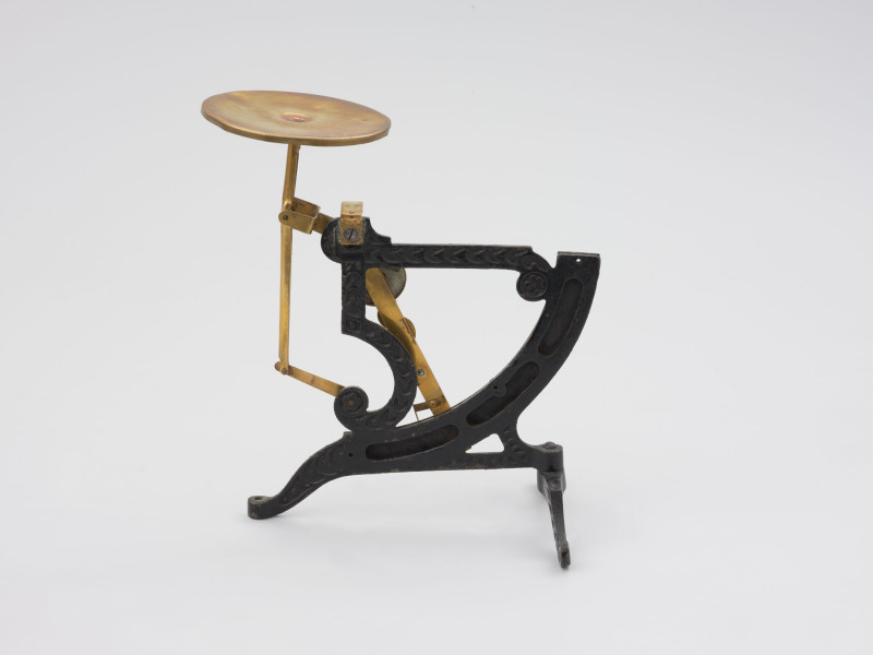 jednowahadłowa uchylna waga pocztowa - Ujęcie z tyłu; Jednowahadłowa uchylna waga pocztowa. Podstawa wagi składa się z dwóch elementów, z których element poprzeczny przymocowany jest do głównego elementu na ruchomo, co pozwala składać nóżkę poprzez jej przekręcenie. Dłuższe ramię podstawy zakończone jest śrubką pozwalającą na regulację wagi w opozycji pionowej. Stalowa konstrukcja łącząca główny trzon wagi z dźwignią nośną nasadzona jest na szalkę.