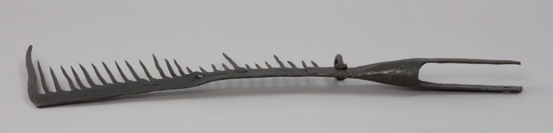 grzebień - Ujęcie z tyłu. 26-zębna ość grzebieniasta, wykonana techniką kowalską. Ramie robocze płaskie, z jednego końca zagięte pod kątem 90 stopni (część ta ma formę zęba grubszego i dłuższego od pozostałych), w drugim końcu przechodzi w tuleję z dwoma pasami, służącą do osadzenia narzędzia na trzonku (trzonka brak). Na końcu każdego pasa otwór – jednego okrągły, drugiego – kwadratowy. Przy nasadzie tulei uszko wykonane z wygiętego w kształcie litery „U” pręta o okrągłym przekroju. Mocowane za pomocą nitu. Zęby mocowane w ramieniu, nierównej długości, ostro zakończone.  Ząb ósmy, licząc od uszka, przynitowany, długi.