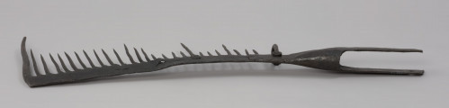 grzebień - Ujęcie z tyłu. 26-zębna ość grzebieniasta, wykonana techniką kowalską. Ramie robocze płaskie, z jednego końca zagięte pod kątem 90 stopni (część ta ma formę zęba grubszego i dłuższego od pozostałych), w drugim końcu przechodzi w tuleję z dwoma pasami, służącą do osadzenia narzędzia na trzonku (trzonka brak). Na końcu każdego pasa otwór – jednego okrągły, drugiego – kwadratowy. Przy nasadzie tulei uszko wykonane z wygiętego w kształcie litery „U” pręta o okrągłym przekroju. Mocowane za pomocą nitu. Zęby mocowane w ramieniu, nierównej długości, ostro zakończone.  Ząb ósmy, licząc od uszka, przynitowany, długi.