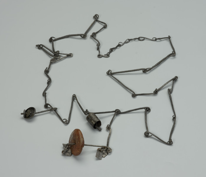 przedmiot ceremonialny, przedmiot religijny i obrzędowy, fetysz - Ujęcie z przodu. Łańcuch składający się z licznych, połączonych ze sobą żelaznych ogniw. Rozdwaja się na końcu tworząc literę Y. Ozdobiony pierścieniami z podwójnym wisiorkiem o trapezowatym kształcie, kamiennym i żelaznym paciorkiem oraz otwartym, żelaznym walcem.