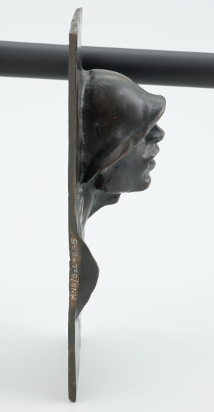 portret męski - Ujęcie z boku z prawej strony; Płaskorzeźba przedstawiająca głowę mężczyzny. Z blachy stanowiącej tło wyłania się kwadratowa twarz z szerokim nosem i rozchylonymi ustami, szyja mocno osadzona, szeroka. Na głowie hutniczy kask. Widoczny fragment torsu.