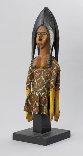 lalka teatralna: Djinè Faro (bóstwo Faro) - Ujęcie z przodu skosem z lewej strony. Drewniana lalka przedstawiająca postać kobiecą o jasnej cerze. Twarz pociągła, nos długi, duże usta, ozdobne nakrycie głowy w kolorze czarnym. Ubrana we wzorzystą sukienkę odsłaniającą nagie piersi.