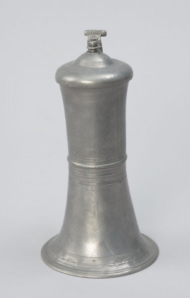 Cynowy dzban o kształcie cylindrycznym - Ujęcie z przodu; Wysmukły dzban o kształcie cylindrycznym z lekko wklęsłym brzuścem, z pierścieniowym zgrubieniem w centrum. Rozszerzająca się ku dołowi podstawa przechodzi w profilowaną stopę. Pokrywa kopulasta, dwustopniowa z profilowaną, toczoną gałką do podnoszenia na szczycie. Na brzuścu u podstawy, pośrodku i przy górnej krawędzi, a także na pokrywie delikatne profilowane i ryte pasy.