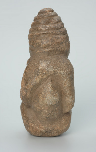 rzeźba; przedmiot obrzędowy; Figura kultu zmarłych - Ujęcie z tyłu. Rzeźbiona w szarobeżowym steatycie postać ludzka ze spiralną linią na czubku dużej głowy, siedząca z przykurczonymi nogami i trzymająca w dłoniach bliżej nieokreślone przedmioty, kształtem przypominające motyki.