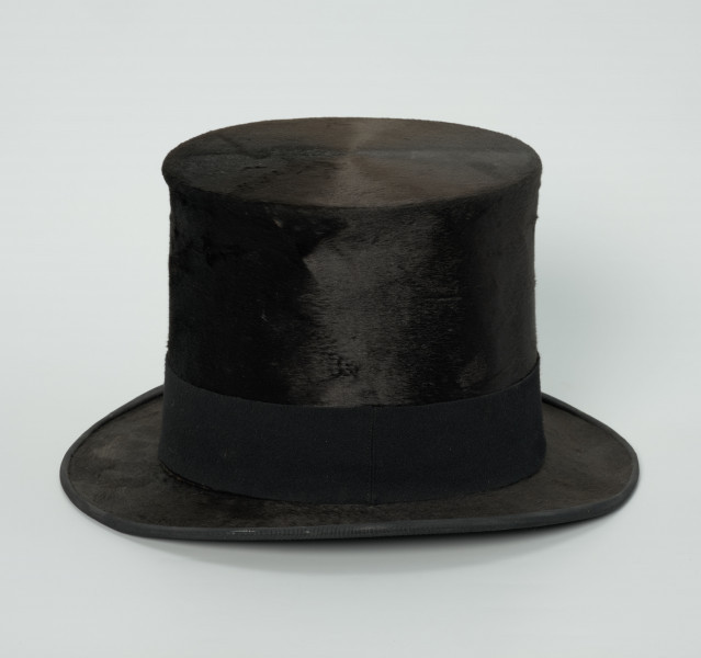 czarny cylinder z welurowej tkaniny - Ujęcie z boku; Czarny cylinder z welurowej tkaniny z czarnym otokiem ponad rondem wykończonym pasmanterią. Wysoki cylinder, sztywny, z główką w kształcie walca oraz małym rondem.