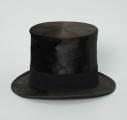 czarny cylinder z welurowej tkaniny - Ujęcie z boku; Czarny cylinder z welurowej tkaniny z czarnym otokiem ponad rondem wykończonym pasmanterią. Wysoki cylinder, sztywny, z główką w kształcie walca oraz małym rondem.