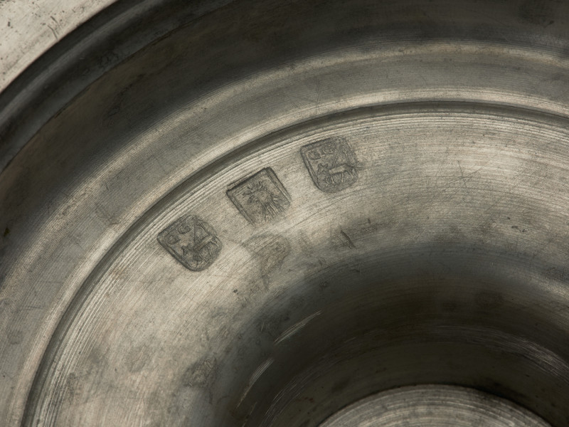 cynowy świecznik na okrągłej, wysklepionej stopie - detal; Stopa świecznika ozdobiona na skraju wypłaszczenia tłoczonym rowkiem, zakończona jest dołem szerokim kręgiem podstawy, tworzącym kryzę z podwójnym tłoczonym rowkiem. Górna część stopy wyniesiona cylindrycznie tworzy oparcie trzonu, złożonego z nakładanych na siebie naprzemiennie pierścieni o dwóch formach: pięciu spłaszczonych kul o obłym wykroju oraz czterech (+ połówka na stopie) nieco niższych o wykroju spiczastym; w połowie wysokości pierścienie zdobi półwałek, zaś górną i dolną nasadę tłoczony rowek. Profitka z szeroką, płaską kryzą ma średnicę zbliżoną do wymiaru stopy. Iglica żelazna, kuta w formie czworobocznego ostrosłupa