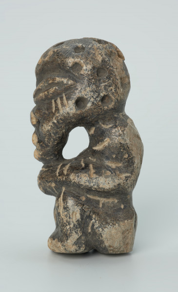 rzeźba; przedmiot obrzędowy; Figura kultu zmarłych - Ujęcie lewy bok. Rzeźbiona w grafitowobeżowym steatycie siedząca postać ludzka, której nieproporcjonalnie dużą głowę zdobią liczne, okrągłe wgłębienia, natomiast ciało pokrywają pionowe i ukośne nacięcia, oznaczające prawdopodobnie skaryfikację. Powierzchnia rzeźby z rysami, mikropęknięciami, przetarciami oraz drobnymi uszkodzeniami.
