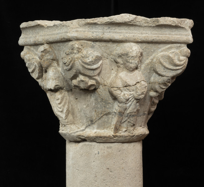 kapitel z przedstawieniem czterech apostołów - Ujęcie trzeciego apostoła z liśćmi po bokach. Kapitel blokowo-kielichowy, rozszerzający się ku górze, wykuty z jednego kawałka kamienia. Okrągłą podstawę otacza wałek o profilu trójkątnym. Zwieńczenie w formie szerokiej czworobocznej płyty abakusa o krawędziach profilowanych wałkiem i wklęską. Na osi każdego boku półplastyczne przedstawienie frontalnie ukazanych postaci Apostołów: Piotra, Pawła, Bartłomieja i Jakuba Starszego. Pomiędzy nimi duże liście palmetowe dekoracyjnie ułożone na narożnikach. Postaci o dużych głowach i dość smukłej budowie, trzymają atrybuty. Długie tuniki i płaszcze o pofałdowanej powierzchni przylegają do sylwetek. Charakterystyczne zdobienie krawędzi szat podwójnym wałkiem i symetrycznie ułożone falujące fryzury. Relief półplastyczny, światłocieniowy. Tło opracowane gładko. Jedno z naroży kapitela uszkodzone. Liczne pęknięcia i ukruszenia na powierzchni kamienia. Pod spodem centralne wgłębienie do umocowania trzpienia łączącego kapitel z trzonem.