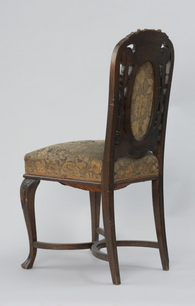 krzesło - Ujęcie z tyłu, z prawej strony. Krzesło w stylu neobarokowym, oparcie i siedzisko tapicerowane. Nogi zwężające się do dołu, przednie wygięte kabriolowo, górne wybrzuszenie ozdobione płaskorzeźbionym motywem palmety, zakończenie dolne trójkątne. Tylne nogi lekko wygięte, powiązane są z przednimi krzyżową poprzeczką umieszczoną u dołu, składającą się z dwóch stykających się w środku półokrągłych pałąkowych listew. Konstrukcja zaplecka stanowi pionowe przedłużenie tylnych nóg o profilowanych krawędziach, zamykających się dwoma półkołami w kształt serca, ponad którym wyprowadzono łukowe zwieńczenie w formie mięsiście rzeźbionego, rozłożonego na boki bukietu liści. Poniżej sercowego styku listew bocznych wydłużony w pionie medalion opasany płaską listwą z wypełnieniem tapicerką. Tkanina w kwiatowy wzór w odcieniach zieleni, różu, żółcieni i ciemnego popielu. Pomiędzy owalem medalionu i listwami oparcia ażurowe wypełnienie mięsistą płaskorzeźbą o motywach roślinnych. Taką samą tkaniną obite jest siedzisko o konstrukcji sprężynowej, stosunkowo wysokie, poszerzające się do przodu, w dolnej części wykończone półwałkiem i pasmanterią, kryjącą krawędź tapicerki.