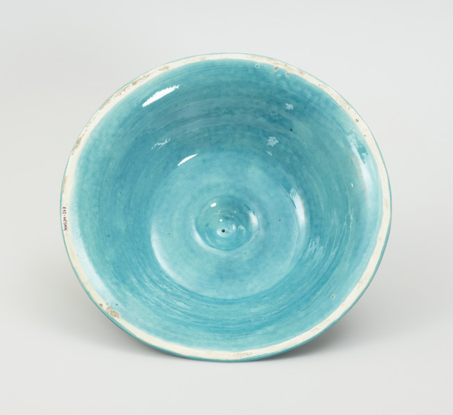 Waza - Ujęcie pokrywki od wnętrza. Waza w kształcie smukłej amfory. Brzusiec ceramiczny, rozszerzający się ku górze, w kolorze kremowym, malowany podszkliwnie (z uzupełnieniami naszkliwnymi) we wzory kwiatowe - bukiety barwnych kwiatów w typie aster w kolorach fioletowo-czerwonym, liliowym, żółtym i białym, z gałązkami i listkami oraz drobnymi kwiatkami przypominającymi bratki o barwie fioletowo-czerwonej i liliowej. Górna część wazy wraz z uchwytami oraz stopą i część podstawy wykonane jako odlewy z mosiądzu. Mosiężna szyja wąska z otokiem perełkowym w środku ozdobiona jest pionowym, rozszerzającym się ku dołowi i ku górze rytmicznym podziałem z ornamentem geometryczno-roślinnym. Na pogrubionym skraju szyi umieszczona jest płytka miseczka w formie kwiatu o wąskich płatkach, stanowiąca wylewkę wazonu. Poniżej dolnej części szyi oprawa wierzchołka brzuśca składająca się z otoku o sercowym wycięciu zwieńczonym profilowaną palmetką oraz dwóch wydłużonych, sięgających 1/3 wysokości brzuśca, pionowych wsporników uchwytów. Te ostatnie składają się z 2 części: zewnętrzna wychodząca ze wsporników opartych na brzuścu, wyniesiona jest po niewielkim skosie do połowy wysokości szyi i na końcu wywinięta do wnętrza wolutowo, a druga - krótsza, skierowana ku dołowi łączy łagodnym łukiem wolutę z górną częścią oprawy brzuśca, kończąc się wywiniętymi na zewnątrz listkami. Uchwyty żłobkowane, zdobione motywem liści. Wazon wsparty jest na mosiężnej podstawie, składającej się z wieńca oplecionego wstęgą, na którym spoczywa zwężająca się ku górze stopa o brzegach ozdobionych perełkowaniem i pierścieniem, wspierającym koszyczek, ujmujący dolną część ceramicznego brzuśca, w formie kwiatu o ostro zakończonych, wąskich płatkach. Całość spoczywa na podstawie z kwadratowej płytki czerwonego marmuru ze ściętymi narożnikami, ujętymi mosiężnymi wspornikami ramy wspierającej płytę od dołu i opartej w narożach na czterech lwich łapach.