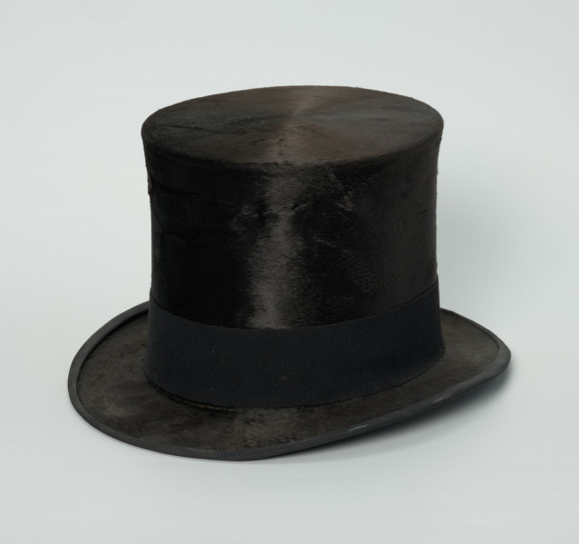 czarny cylinder z welurowej tkaniny - Ujęcie ze skosu; Czarny cylinder z welurowej tkaniny z czarnym otokiem ponad rondem wykończonym pasmanterią. Wysoki cylinder, sztywny, z główką w kształcie walca oraz małym rondem.