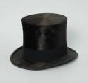 czarny cylinder z welurowej tkaniny - Ujęcie ze skosu; Czarny cylinder z welurowej tkaniny z czarnym otokiem ponad rondem wykończonym pasmanterią. Wysoki cylinder, sztywny, z główką w kształcie walca oraz małym rondem.