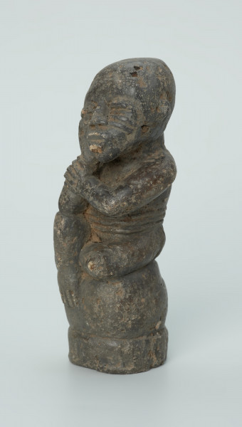figura kultu przodków - Ujęcie z przodu z lewej. Rzeźbiona w grafitowym kamieniu postać ludzka w pozycji siedzącej - najprawdopodobniej mężczyzny. Postać siedzi na wysokiej podstawie, która swym kształtem przypomina kamień. Rzeźba ma charakterystyczne poziome nacięcia widoczne na obu policzkach. Widoczne rysy, mikropęknięcia i liczne malutkie otworki.