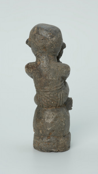 figura kultu przodków - Ujęcie z tyłu. Rzeźbiona w grafitowym kamieniu postać ludzka w pozycji siedzącej - najprawdopodobniej mężczyzny. Postać siedzi na wysokiej podstawie, która swym kształtem przypomina kamień. Rzeźba ma charakterystyczne poziome nacięcia widoczne na obu policzkach. Widoczne rysy, mikropęknięcia i liczne malutkie otworki.