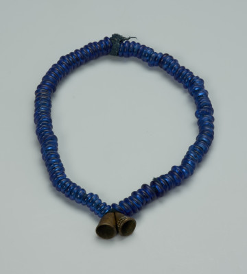naszyjnik , biżuteria , ozdoba - Ujęcie z przodu. Zamknięty, kobaltowy naszyjnik. Na bawełniany sznurek nałożone są szklane, kobaltowe paciorki oraz wykonane z brązu dwa wisiorki w kształcie stożków przypominające dzwonki. Wisiorki ozdobiono nacięciami.