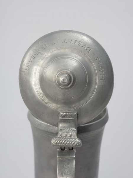 Cynowy dzban o kształcie cylindrycznym - detal górnej części; Fragment górnej części dzbana. Zawias podtrzymujący pokrywę. Pokrywa kopulasta, dwustopniowa z profilowaną, toczoną gałką do podnoszenia na szczycie.